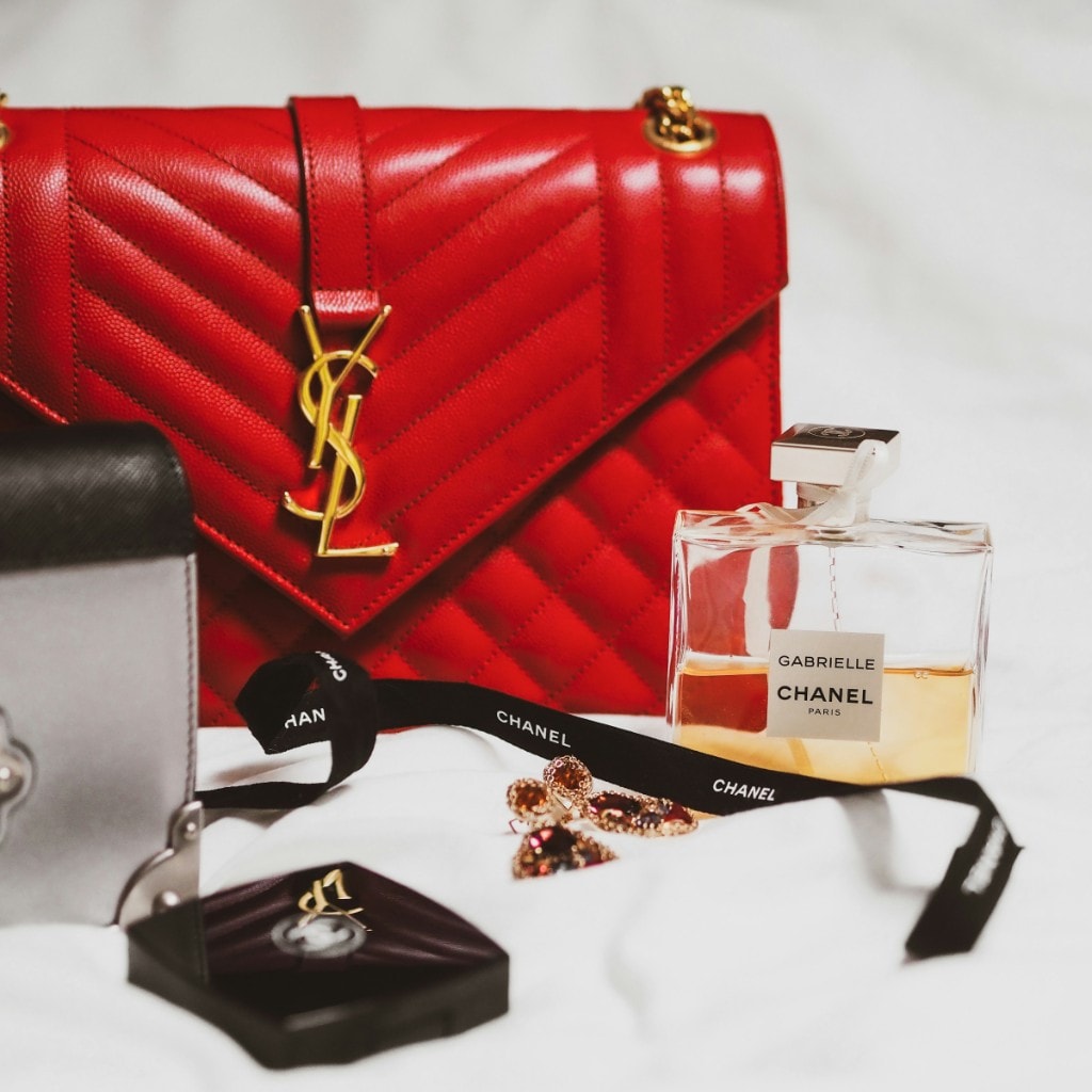 A red Saint Laurent envelope bag styled with a Chanel Gabrielle fragrance and Chanel ribbon on a soft white backdrop.